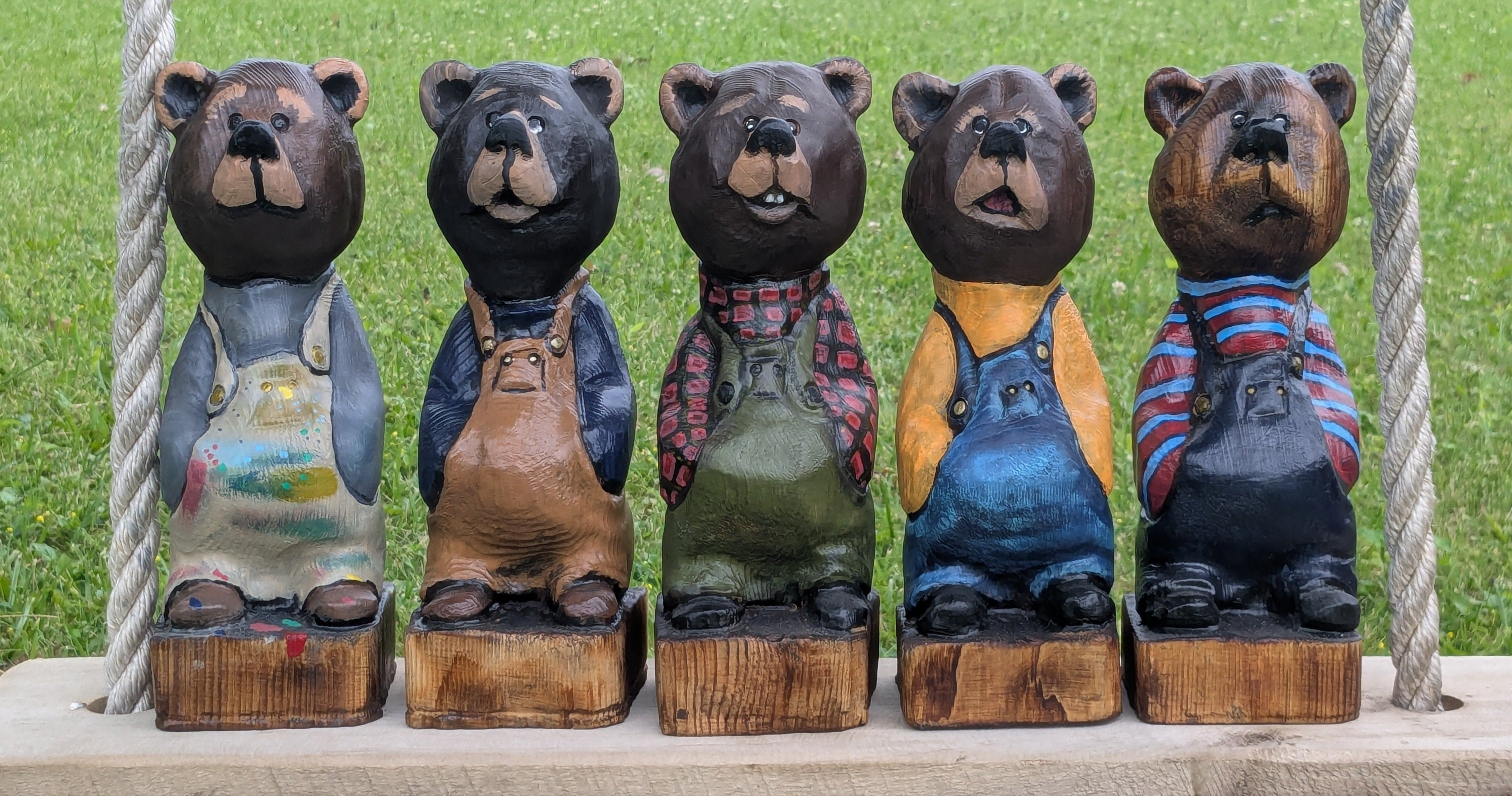 Five hand-crafted wooden bear figurines wearing colorful overalls and work clothes, displayed side-by-side on a wooden platform with rope supports against a green grassy park background.